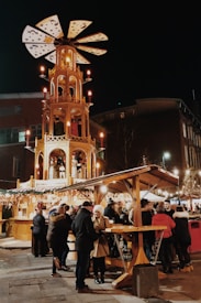 A bustling outdoor market scene at night with a large, illuminated wooden structure resembling a traditional Christmas pyramid in the center. People are gathered around wooden stalls decorated with festive lights, engaging in lively conversations and enjoying the atmosphere.