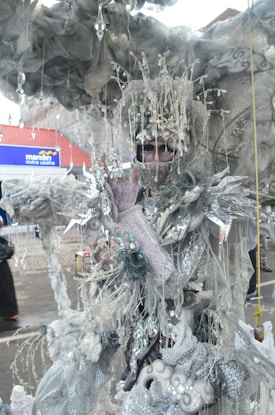 A person is wearing an elaborate, fantastical costume adorned with intricate details such as shimmering beads, crystal-like ornaments, and wispy, cloud-like structures. The costume features a metallic and ethereal color palette, creating an imaginative and otherworldly effect.