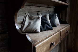 A rustic wooden shelf with a series of small fabric bags in various shades of beige and gray. The shelf features drawers with round wooden knobs, set against a backdrop of weathered wooden planks.