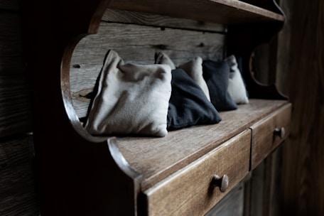 A rustic wooden shelf with a series of small fabric bags in various shades of beige and gray. The shelf features drawers with round wooden knobs, set against a backdrop of weathered wooden planks.