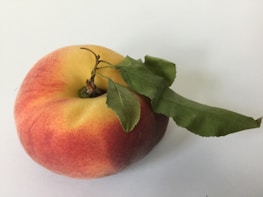 A ripe peach with a smooth gradient of red, orange, and yellow hues sits on a light surface. Two green leaves are attached to the stem, adding a fresh and natural element to the composition.