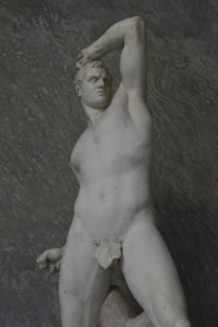 A classical marble statue depicting a muscular male figure in a powerful pose. The figure stands with one arm raised and bent over the head, and the other arm rests on the hip. The sculpture captures intricate details of the human form, showcasing defined muscles and a strong facial expression.