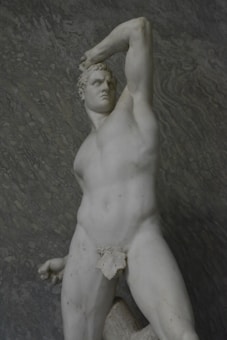 A classical marble statue depicting a muscular male figure in a powerful pose. The figure stands with one arm raised and bent over the head, and the other arm rests on the hip. The sculpture captures intricate details of the human form, showcasing defined muscles and a strong facial expression.