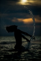 A silhouette of a person with long hair is captured against the backdrop of a setting or rising sun, creating a dramatic contrast. Water splashes around them in an elegant arc, suggesting movement and energy. The scene conveys a sense of freedom and connection with nature.