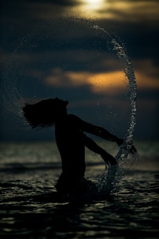 A silhouette of a person with long hair is captured against the backdrop of a setting or rising sun, creating a dramatic contrast. Water splashes around them in an elegant arc, suggesting movement and energy. The scene conveys a sense of freedom and connection with nature.