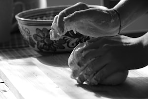 Hands are kneading dough on a wooden surface, with a patterned bowl in the background. The scene captures a moment of cooking or baking, showcasing the tactile engagement with the dough.