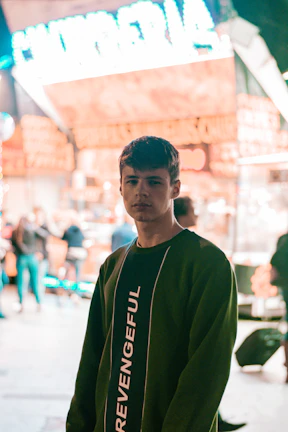 A young man is looking into the camera while wearing a dark sweatshirt with the word 'REVENGEFUL' printed on it. He stands in front of a brightly lit storefront with neon signs and blurry figures of people in the background.
