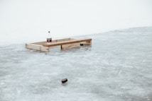 A wooden frame sits on an icy surface with a bottle placed on top. In the foreground, a hockey puck is visible on the ice.