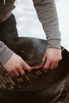 Hands placing meat onto a charcoal grill, with smoke lightly rising from the coals. The person is wearing a gray long-sleeve shirt. The setting appears to be outdoors.