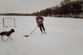 A person in a hockey jersey is playing ice hockey on a frozen lake with a dog wearing a sweater. There is a hockey net behind them, and the surrounding landscape includes trees and snow.