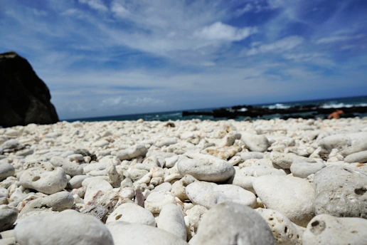 A serene rocky beach with smooth black and gray stones under a bright blue sky dotted with fluffy white clouds.