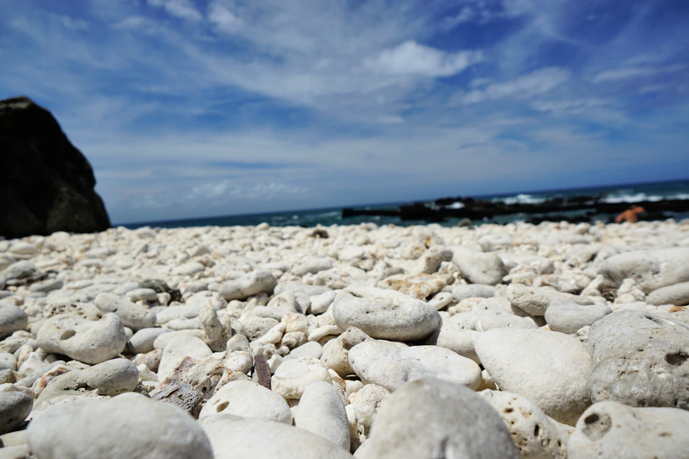 A serene rocky beach with smooth black and gray stones beside calm, clear blue water under a sky dotted with soft white clouds.