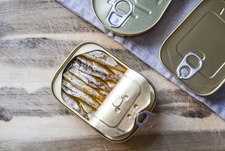 Three rectangular tin cans with pull tab openers, two of which are closed and one opened, reveal neatly packed fish (likely sardines). These tin cans are placed on a wooden surface partially covered by a grey cloth, providing a rustic setting.