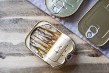 Three rectangular tin cans with pull tab openers, two of which are closed and one opened, reveal neatly packed fish (likely sardines). These tin cans are placed on a wooden surface partially covered by a grey cloth, providing a rustic setting.