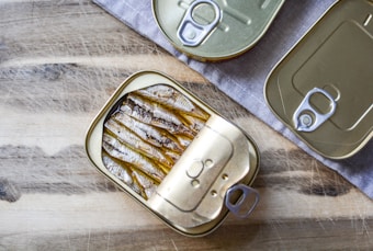 Three rectangular tin cans with pull tab openers, two of which are closed and one opened, reveal neatly packed fish (likely sardines). These tin cans are placed on a wooden surface partially covered by a grey cloth, providing a rustic setting.