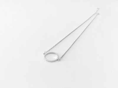 A delicate silver necklace with a thin chain and a circular pendant lying on a white surface. The pendant is smooth and appears to be made of metal, matching the material of the chain.
