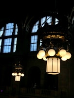 Large, ornate chandeliers with glowing bulbs hang from the high ceiling of a grand interior space. Tall windows with multiple panes let in natural light, contrasting the illuminated fixtures.