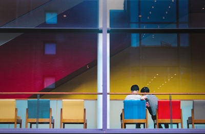 Two people sit closely together on colorful chairs, back to back, facing a large glass window. A row of chairs in various colors including yellow, blue, red, and green is aligned against the window. In the background, a vibrant wall with red, yellow, and blue sections is visible. The setting appears to be indoors with modern architectural design visible through the window.
