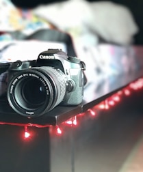 A DSLR camera is positioned on the edge of a dark surface, surrounded by a strand of red fairy lights. The background is softly blurred, suggesting an indoor setting with various colors.