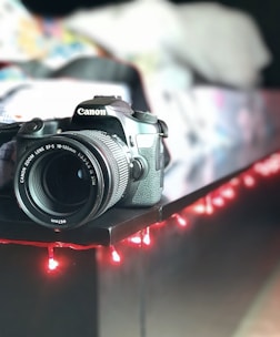 A DSLR camera is positioned on the edge of a dark surface, surrounded by a strand of red fairy lights. The background is softly blurred, suggesting an indoor setting with various colors.
