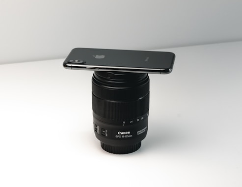 A smartphone is placed horizontally on top of a Canon camera lens resting on a flat surface. The setup is minimalist and well-lit, creating a sense of balance between the two objects.