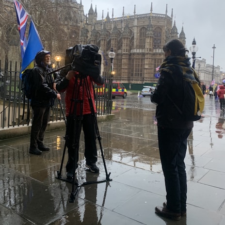 A rainy street scene featuring a camera crew filming an individual, with a historic Gothic-style building in the background. People carry blue and red flags, and vehicles are parked nearby.
