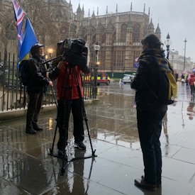 A rainy street scene featuring a camera crew filming an individual, with a historic Gothic-style building in the background. People carry blue and red flags, and vehicles are parked nearby.