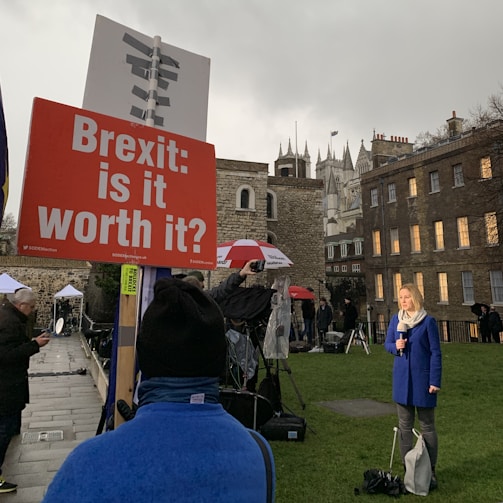 A reporter is standing in a park holding a microphone, with a large sign in the foreground reading 'Brexit: is it worth it?'. There are several people around, including someone holding an umbrella, and a backdrop of historic buildings under overcast skies.