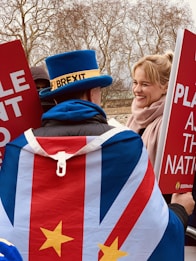 A person wearing a distinctive blue top hat with a label and a flag-themed cape stands amidst a political protest. They interact with another person who is smiling, both holding large red signs with white text. The background features leafless trees indicating a cold or winter season.