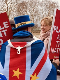A person wearing a distinctive blue top hat with a label and a flag-themed cape stands amidst a political protest. They interact with another person who is smiling, both holding large red signs with white text. The background features leafless trees indicating a cold or winter season.