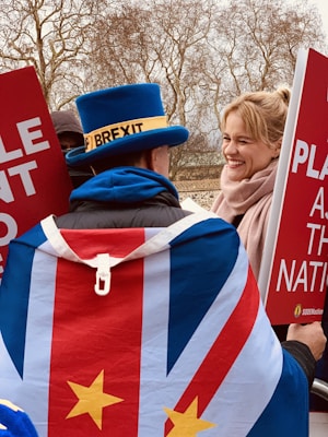 A person wearing a distinctive blue top hat with a label and a flag-themed cape stands amidst a political protest. They interact with another person who is smiling, both holding large red signs with white text. The background features leafless trees indicating a cold or winter season.