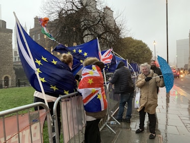 Several people gather outside, holding EU and UK flags, standing near metal barricades on a rainy day. One person struggles with a blue polka dot umbrella, and others are wrapped in flags as they brace against the wind.