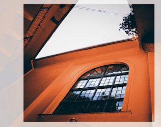 A perspective view of an architectural structure with an arched window and grid-like panes framed by orange-colored walls. The view includes an outdoor angle with a portion of a building and some greenery visible against a clear sky.