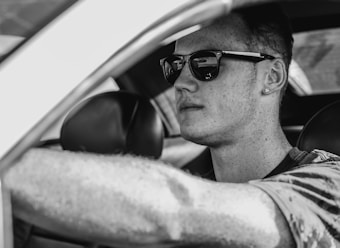 A person wearing sunglasses is sitting in the driver's seat of a car. The image is in black and white, which adds a classic and cool vibe. The individual appears focused and has short hair, with a visible earring. The interior of the car is slightly visible, emphasizing the driver's side and steering wheel.