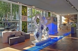 A modern showroom with large windows allowing natural light to illuminate the space. The interior features a geometric sculpture with reflective surfaces, set on a polished wooden floor. Greenery adorns the walls, adding a touch of nature. Flat-screen monitors display images of a car, and there's seating made of dark gray fabric. The atmosphere is sleek and futuristic.