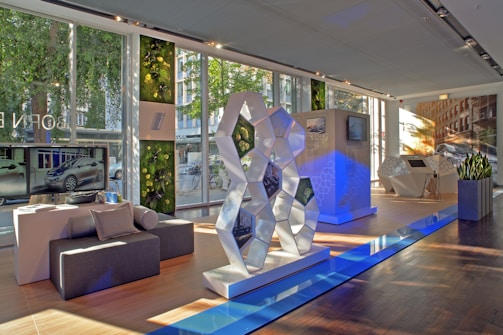 A modern showroom with large windows allowing natural light to illuminate the space. The interior features a geometric sculpture with reflective surfaces, set on a polished wooden floor. Greenery adorns the walls, adding a touch of nature. Flat-screen monitors display images of a car, and there's seating made of dark gray fabric. The atmosphere is sleek and futuristic.