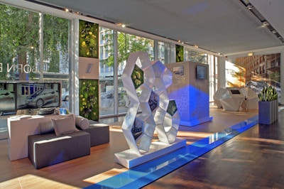 A modern showroom with large windows allowing natural light to illuminate the space. The interior features a geometric sculpture with reflective surfaces, set on a polished wooden floor. Greenery adorns the walls, adding a touch of nature. Flat-screen monitors display images of a car, and there's seating made of dark gray fabric. The atmosphere is sleek and futuristic.