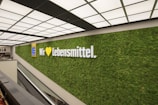 A large, well-lit wall with a lush green plant covering displays a prominent logo and the words 'Wir lieben Lebensmittel'. The setting is modern and minimalist, featuring a ceiling with a grid of lights and an escalator nearby.