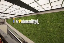 A large, well-lit wall with a lush green plant covering displays a prominent logo and the words 'Wir lieben Lebensmittel'. The setting is modern and minimalist, featuring a ceiling with a grid of lights and an escalator nearby.