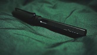 A black pen rests on a textured green fabric. The focus is on the sleek design of the pen, highlighting its modern and minimalist features. The lighting creates subtle shadows and brings out the richness of the green background.