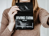 A person holding a bag of Irving Farm New York coffee in front of their chest, with visible text describing the coffee as a medium roast with notes of milk chocolate and clementine. The person has long blonde hair and is wearing a brown sweater.
