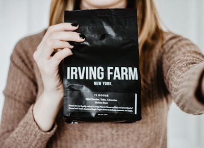 A person holding a bag of Irving Farm New York coffee in front of their chest, with visible text describing the coffee as a medium roast with notes of milk chocolate and clementine. The person has long blonde hair and is wearing a brown sweater.