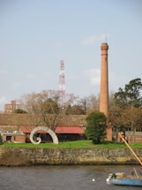 A tall brick chimney stands prominently next to a building with a red roof. In front of the building is a spiral-shaped sculpture. The area is surrounded by trees, some of which have bare branches, possibly indicating a winter or autumn setting. A water body is visible in the foreground, with stones forming a retaining wall.