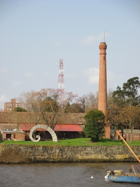 A tall brick chimney stands prominently next to a building with a red roof. In front of the building is a spiral-shaped sculpture. The area is surrounded by trees, some of which have bare branches, possibly indicating a winter or autumn setting. A water body is visible in the foreground, with stones forming a retaining wall.