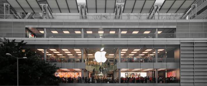 A multi-level, modern retail store with a large glowing logo on the exterior. The store features wide glass windows and numerous people inside, suggesting a bustling atmosphere. The building's architecture includes clean lines and a symmetrical design, integrating greenery outside.