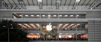 A multi-level, modern retail store with a large glowing logo on the exterior. The store features wide glass windows and numerous people inside, suggesting a bustling atmosphere. The building's architecture includes clean lines and a symmetrical design, integrating greenery outside.