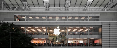 A multi-level, modern retail store with a large glowing logo on the exterior. The store features wide glass windows and numerous people inside, suggesting a bustling atmosphere. The building's architecture includes clean lines and a symmetrical design, integrating greenery outside.
