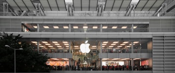 A multi-level, modern retail store with a large glowing logo on the exterior. The store features wide glass windows and numerous people inside, suggesting a bustling atmosphere. The building's architecture includes clean lines and a symmetrical design, integrating greenery outside.