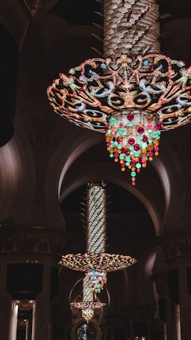 Large, ornate chandeliers hang from the ceiling in an opulent interior space, showcasing intricate designs with bright colored crystals and lights, creating a luxurious atmosphere with architectural arches in the background.
