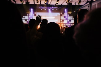A dimly lit indoor venue with a crowd of people facing a stage. Warm lighting and a large screen are visible in the background. One person has their hand raised, suggesting engagement or participation.